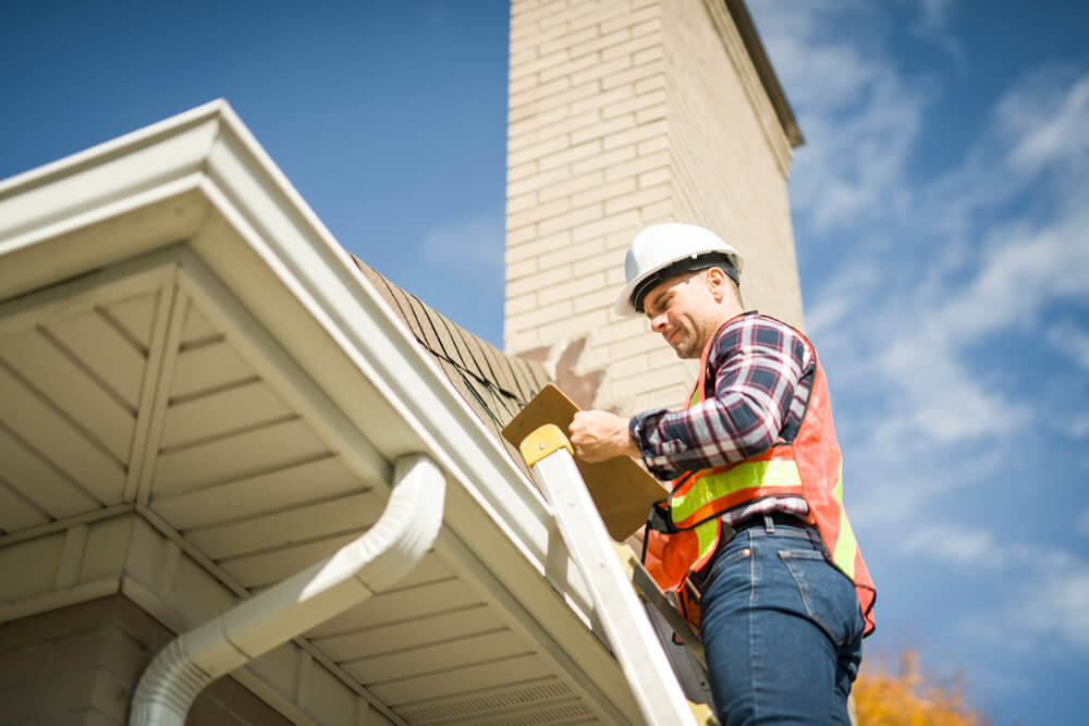 A roofing contractor standing on a ladder inspecting a roof and making notes