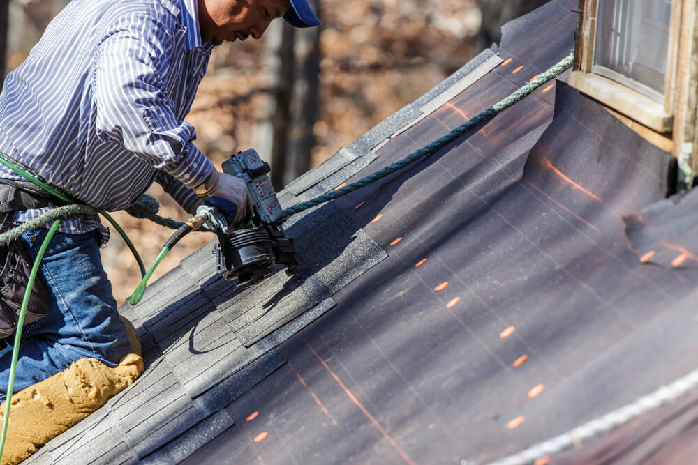 Roofing worker kneeling on a rooftop, using a nail gun to nail shingles to a roof