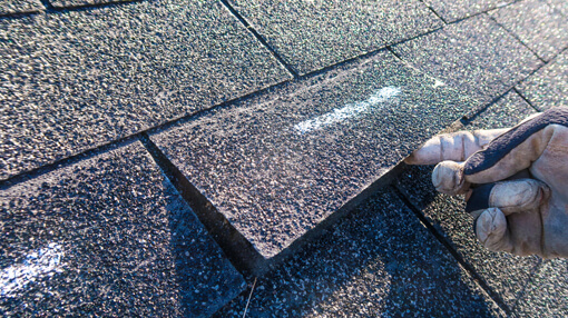 A close up of a roof inspector's hand lifting a shingle and marking it with white chalk