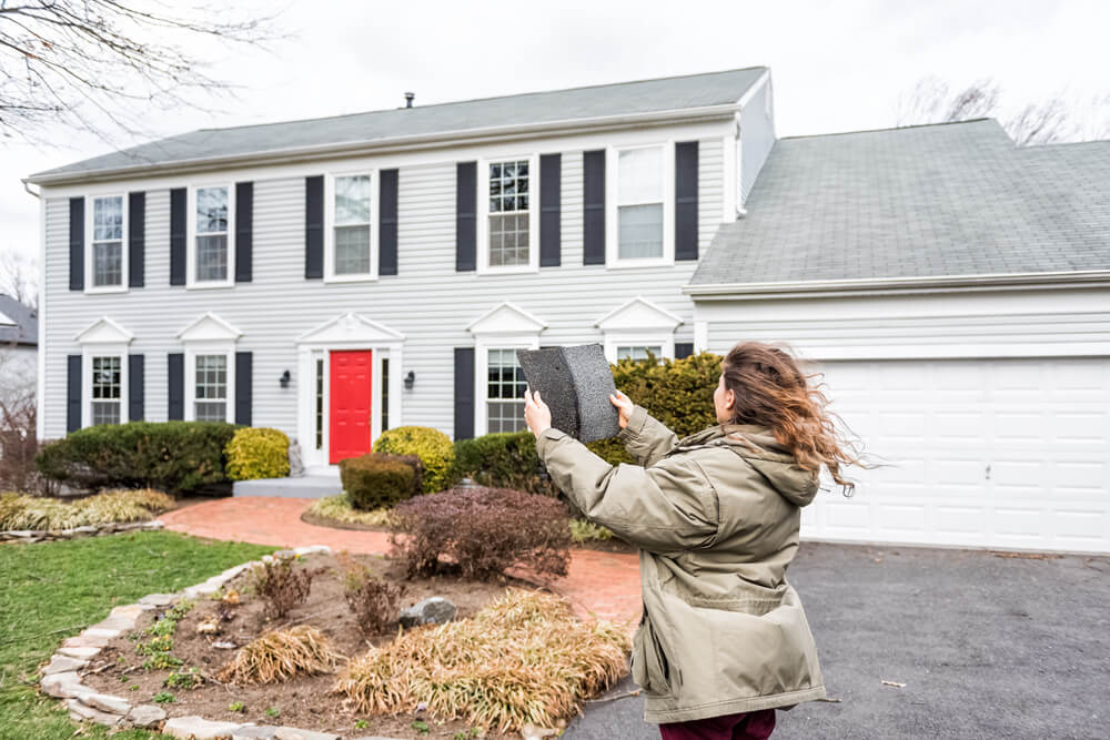 Woman standing in front of her home, holding up 2 different color roofing shingles, trying to decide which one to pick